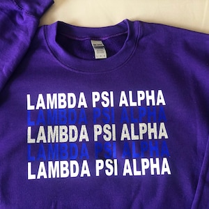 May include: Purple sweatshirt with the text "Lambda Psi Alpha" repeated five times in white letters with a blue glitter effect.