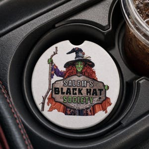 May include: A white car coaster featuring a green-skinned witch with red hair and a black hat, holding a wooden sign that reads "Salem's Black Hat Society." The coaster is in a car cup holder.
