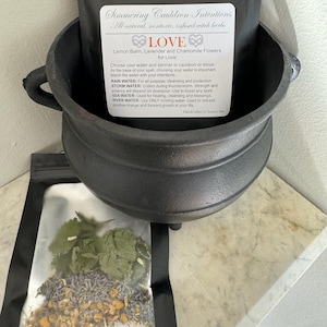 May include: A black cauldron with a white label that reads "Simmering Cauldron Intentions All natural, nontoxic, infused witch herbs LOVE Lemon Balm, Lavender and Chamomile Flowers for Love Choose your water and simmer in cauldron or stove. As the base of your spell, choosing your water is important. Match the water with your intentions. RAIN WATER: For all purpose, cleansing and protection STORM WATER: Collect during thunderstorm. Strength and potency will depend on downpour. Use to boost any spell SEA WATER: Used for healing, cleansing and blessings. RIVER WATER: Use ONLY running water. Used to release positive change and forward growth in your life Handcrafted in Salem, MA". A clear plastic bag with dried herbs inside is sitting in the cauldron.