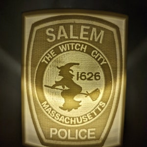 Puede incluir: Una luz nocturna beige con forma de placa policial con el texto "SALEM THE WITCH CITY 1626 MASSACHUSETTS POLICE" en blanco.
