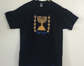 May include: A black t-shirt with a blue trim and fringe at the bottom. The shirt features a gold embroidered image of the Star of David, a menorah, and the Hebrew word "Yisrael" (Israel).