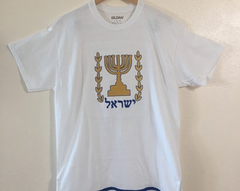 May include: White t-shirt with a gold menorah and olive branches design. The Hebrew word "ישראל" is printed below the menorah. The shirt has a blue and white fringe trim.
