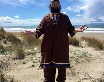 May include: A man wearing a brown robe with gold embroidery and a brown hat stands with his arms outstretched on a sandy beach. The robe has a blue and white stripe at the bottom. The sky is blue with white clouds.