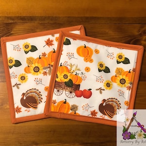 May include: Two orange and white fabric pot holders with a Thanksgiving theme. The pot holders feature a pattern of pumpkins, sunflowers, turkeys, and a wagon full of pumpkins. The pot holders have a brown border.