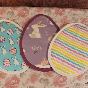 May include: Three colorful Easter-themed pot holders in the shape of eggs. One pot holder is blue with white bunnies and colorful eggs, another is purple with a white bunny and floral accents, and the last is white with pastel stripes.