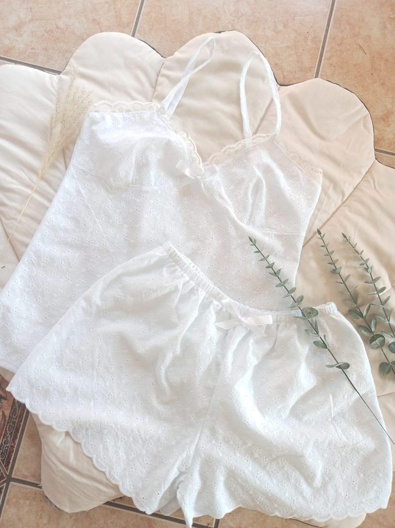 May include: White eyelet camisole and shorts set. The camisole has spaghetti straps and a scalloped lace trim. The shorts have an elastic waistband and a small bow. The set is made of a lightweight, breathable fabric.