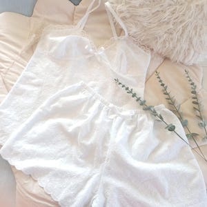 May include: White eyelet camisole and shorts set. The camisole has spaghetti straps and a small bow. The shorts have a scalloped hem. The set is made of a soft, lightweight fabric. A fluffy pillow and sprigs of greenery are in the background.
