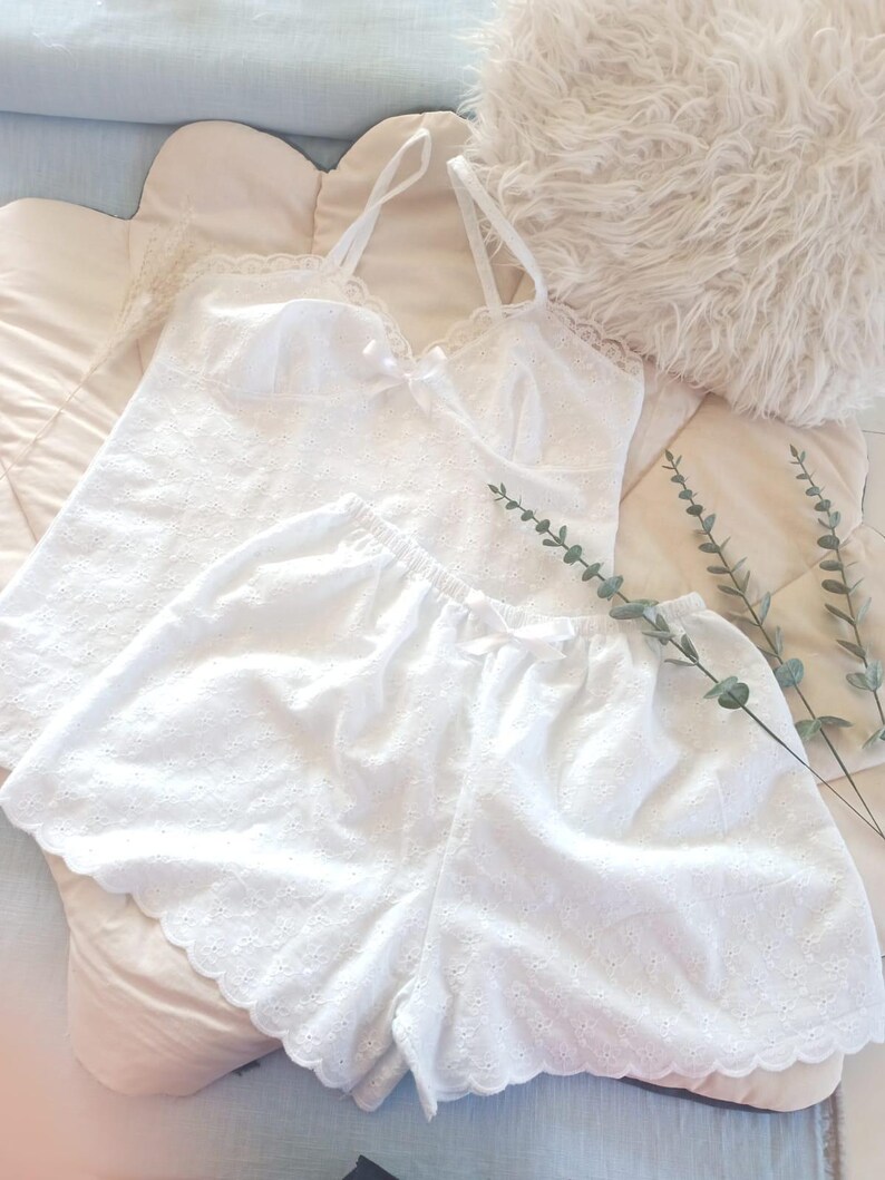 May include: White eyelet camisole and shorts set. The camisole has spaghetti straps and a small bow. The shorts have a scalloped hem. The set is made of a soft, lightweight fabric. A fluffy pillow and sprigs of greenery are in the background.