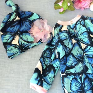 Puede incluir: Un conjunto de ropa para bebé con estampado de mariposas azules y blancas. El conjunto incluye un mono de manga larga y un gorro anudado con una flor rosa.