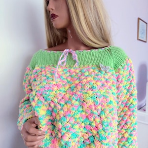May include: A colorful, cropped sweater with a textured knit pattern. The sweater features a bright green neckline and a tie detail. The body of the sweater is a mix of pastel colors, including pink, yellow, and blue. A silver brooch is attached to the sweater.