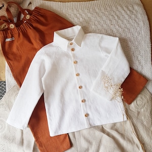 May include: A white linen button-down shirt with long sleeves and a brown fabric background. The shirt has a collar and six buttons down the front.