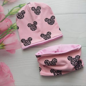 Może przedstawiać: Różowy zestaw dla dziecka: czapka i ocieplacz na szyję. Zestaw posiada powtarzający się wzór czarnych sylwetek głowy Myszki Miki w panterkę. Czapka i ocieplacz wykonane są z miękkiej, różowej tkaniny.
