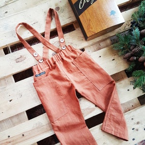 Può includere: Pantaloni in lino color ruggine con bretelle e due grandi tasche. I pantaloni hanno un'etichetta che dice "Exclusive".