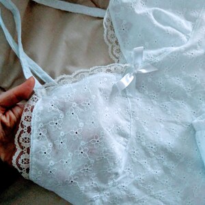 May include: A light blue eyelet fabric camisole with delicate floral embroidery. The camisole features thin straps, a lace trim, and a small white bow. The fabric appears soft and lightweight, suitable for sleepwear or layering.