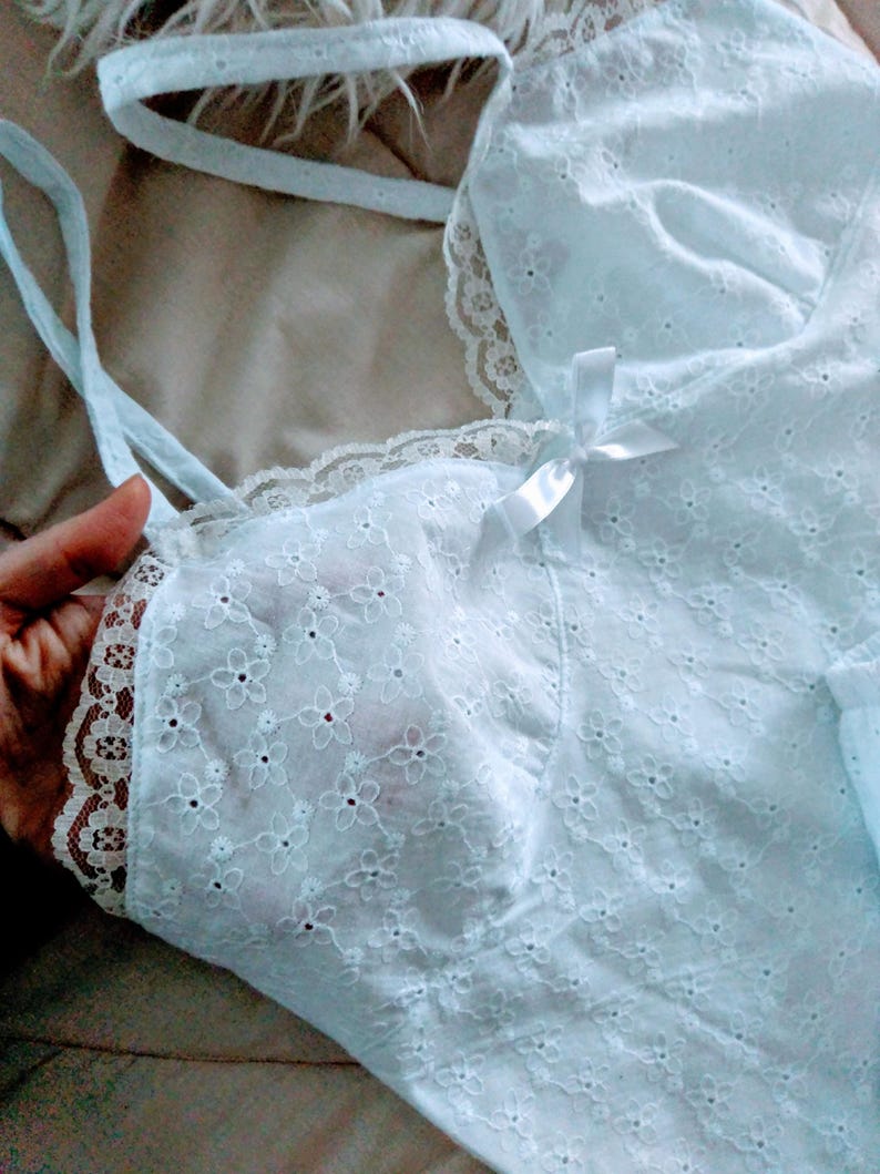 May include: A light blue eyelet fabric camisole with delicate floral embroidery. The camisole features thin straps, a lace trim, and a small white bow. The fabric appears soft and lightweight, suitable for sleepwear or layering.