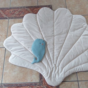 May include: A white, plush, quilted play mat shaped like a seashell with a light blue whale toy in the center.
