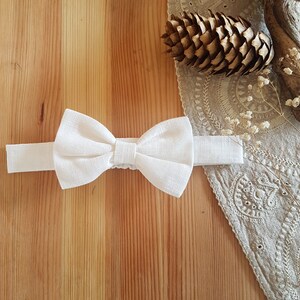 May include: A white linen bow tie with a simple design. The bow tie is on a wooden surface.