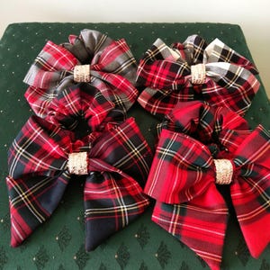 May include: Six tartan fabric hair bows in red, black, grey, and white. Each bow has a decorative centre piece. The bows are arranged on a green surface with a diamond pattern.