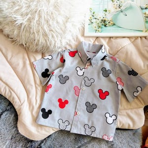 Könnte beinhalten: Ein graues, kurzärmeliges Hemd mit Knöpfen und einem Mickey-Mouse-Druck. Das Hemd hat schwarze, weiße und rote Mickey-Mouse-Köpfe auf grauem Hintergrund.