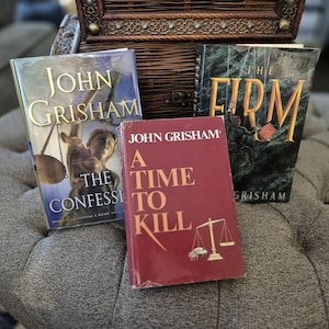 May include: Three John Grisham novels: "The Confession," "A Time to Kill," and "The Firm." The books are on a gray upholstered surface, with a decorative wooden chest in the background. The book covers feature text and illustrations.