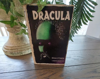 Puede incluir: Una copia vintage del libro "Drácula" de Bram Stoker. La portada presenta una ilustración de un rostro verde con ojos morados sobre un fondo negro. El título "DRACULA" está en blanco, con el nombre del autor y "A Modern Library Book" debajo.