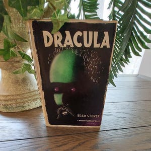May include: A hardcover book titled Dracula by Bram Stoker, published by Modern Library. The cover features a dark, stylized illustration of a face with a green forehead, purple eyes, and visible fangs against a black background with white text.