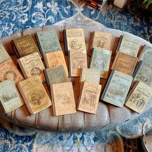 May include: A collection of twenty-two antique hardcover schoolbooks with weathered cloth and paper covers in shades of tan, brown, green, and gray. The books feature various titles including Arithmetic, Readers, and Spellers, with visible text and illustrations.