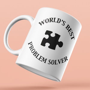 Puede incluir: Taza de cerámica blanca con un gráfico negro de una pieza de rompecabezas y el texto "World's Best Problem Solver".