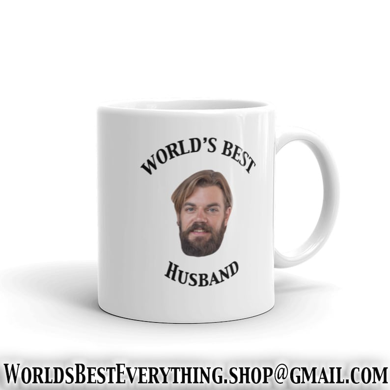 May include: White ceramic mug with a black inscription that reads "World's Best Husband". A man's face with a beard is in the center of the mug.
