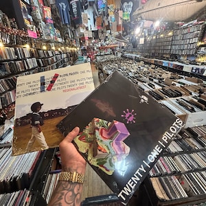 May include: Two vinyl records by Twenty One Pilots are held in a record store. One album cover features a person walking, the other has a black background with colorful abstract shapes and the band's name. The store is filled with records and band merchandise.
