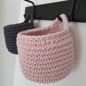 May include: Two crocheted hanging baskets, one pink and one gray, are hanging on a black metal hook.