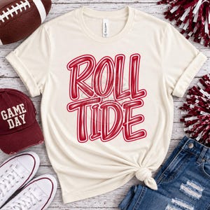 May include: A cream-colored short-sleeve t-shirt featuring the red text ROLL TIDE in a bold, stylized font. The shirt has a crew neckline and a knotted hem detail at the bottom, showcasing a casual design suitable for sports-themed apparel.