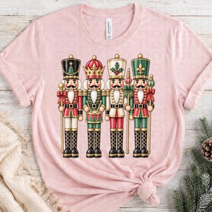 May include: A light pink short-sleeve t-shirt featuring a graphic print of four nutcracker figures standing in a row. The nutcrackers are dressed in red, green, and white uniforms with gold accents, each wearing a distinct hat and holding a staff.