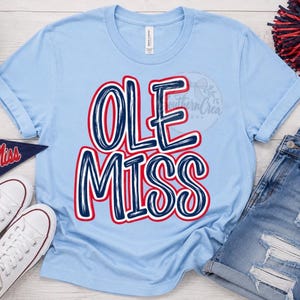 May include: A light blue short-sleeved t-shirt featuring the text OLE MISS in large, bold, navy blue letters with a thick red outline. The lettering has a textured, hand-drawn appearance. The shirt has a crew neckline and a standard fit.