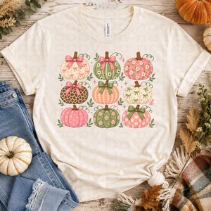 以下が含まれることがあります： クリーム色の半袖Tシャツで、9つのスタイリッシュなカボチャがグリッド状に並んだグラフィックプリントが施されています。それぞれのカボチャには、花柄、水玉模様、ヒョウ柄、チェック柄などのユニークなパターンがあり、リボンで装飾されています。