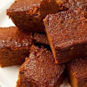 May include: A close-up of a plate of freshly baked gingerbread squares. The squares are a rich brown colour and have a glossy glaze.