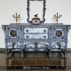 May include: A vintage gray and silver painted buffet with ornate carvings and gold accents. The buffet has two drawers and two doors. It is sitting on a dark tiled floor. The text "HDS Hawthorne Design Studio" is visible in the bottom right corner of the image.