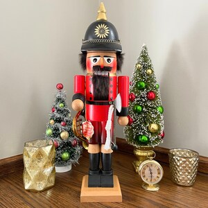 May include: A wooden nutcracker figurine dressed as a soldier, wearing a red and black uniform with a gold trim, holding a musical instrument. The nutcracker is standing on a wooden base and is surrounded by small Christmas trees.