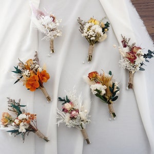 Op de afbeelding: Een assortiment van zeven corsages met gedroogde bloemen in tinten oranje, wit en groen. Elke corsage is gewikkeld met touw en versierd met kleine parels. De corsages zijn geplaatst op een witte stoffen achtergrond.