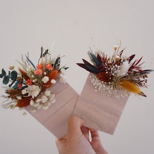 May include: Two small, pink fabric envelopes, each decorated with a vibrant arrangement of dried flowers. The floral arrangements feature orange, white, and burgundy blooms, with accents of green and yellow. The envelopes are held up against a neutral background.