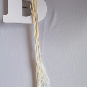 Puede incluir: Una letra "B" de madera blanca con un manojo de tallos secos de color crema y plumas. Los tallos son largos y delgados, con las plumas en la parte inferior. El fondo es una tela gris claro.