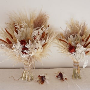 Op de afbeelding: Twee boeketten en twee corsages van gedroogde bloemen. De boeketten bevatten pampasgras, rode rozen en witte vulbloemen. De corsages hebben vergelijkbare elementen. De boeketten zijn gebonden met touw en lint.