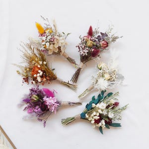 Pode incluir: Uma coleção de pequenos bouquets de flores secas em várias cores, incluindo amarelo, laranja, rosa, roxo e branco. Cada bouquet é amarrado com barbante e repousa sobre uma superfície branca. Os bouquets apresentam uma variedade de texturas e formas.