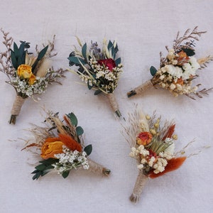 Op de afbeelding: Vijf droogbloem corsages. Elke corsage heeft een verscheidenheid aan droogbloemen in tinten oranje, geel, wit en groen, gewikkeld met touw. De corsages zijn gerangschikt op een neutraal gekleurde stoffen achtergrond.