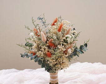 May include: A dried flower bouquet featuring a variety of textures and colors. The arrangement includes white, orange, and green elements, with a burlap and lace-wrapped stem. The bouquet rests on a pink fabric.