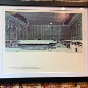 May include: A framed print featuring a detailed illustration of a circular spacecraft inside a large industrial hangar. The artwork includes text at the bottom describing the scene as a Death Star hangar. The frame is black with a white mat border.