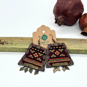 Könnte beinhalten: Paar weinrote Stoffohrringe mit floraler Stickerei in Rot, Grün und Gelb. Die Ohrringe haben eine rechteckige Form mit dekorativen Metallbesatz und hängenden Metallakzenten. Die Ohrringe werden mit einem Etikett mit der Aufschrift "BOHE ME BERUJA" präsentiert.