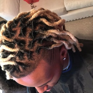 May include: A person with black and blonde dreadlocks. The dreadlocks are styled in a way that covers most of the head. The person is wearing a black shirt.