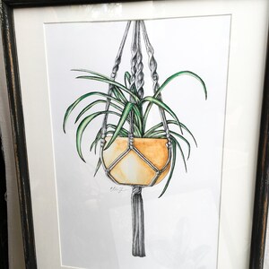 May include: A watercolor illustration of a potted plant hanging from a macrame plant hanger. The plant is green and the pot is a light orange color. The hanger is gray and has a tassel at the bottom. The illustration is signed "ellenj" in the bottom left corner.