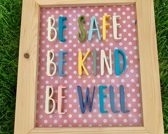 May include: A wooden framed sign with a pink polka dot background and the words "Be Safe Be Kind Be Well" in colorful letters.